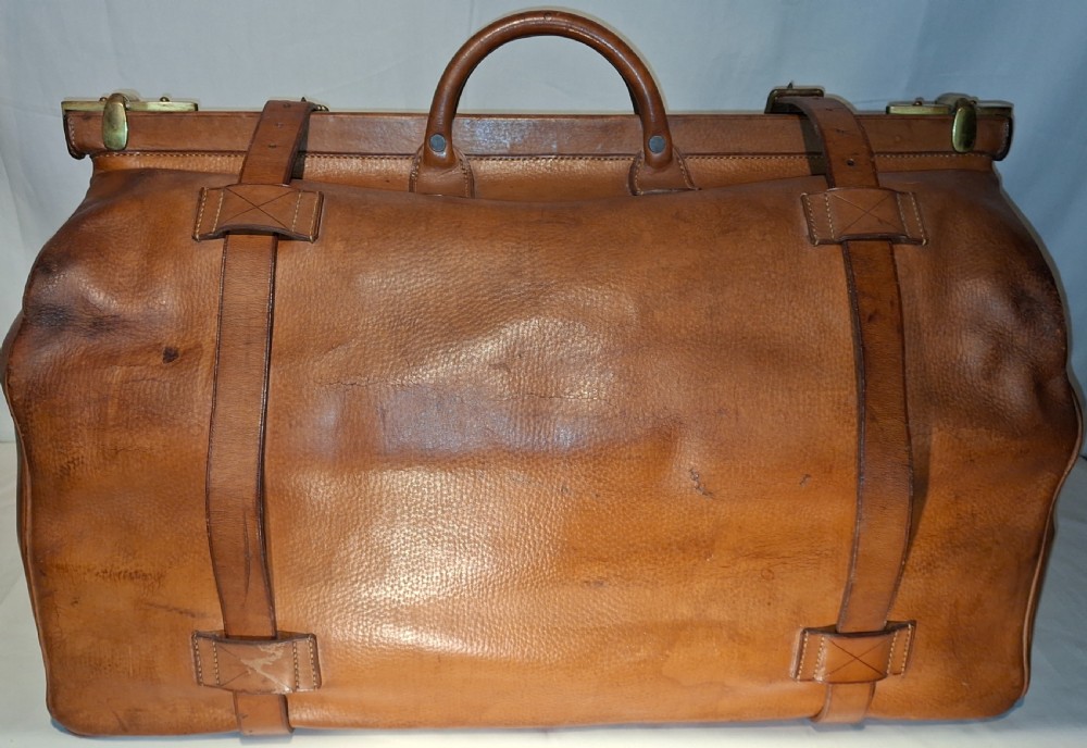 A warm brown leather Gladstone bag featuring twin buckle straps, a brass central lock mechanism, a thick handle, and gold initials. View IV