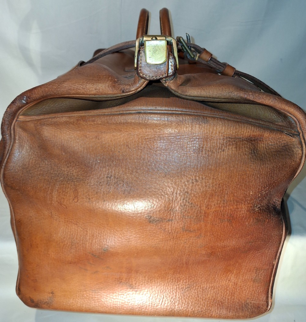 A warm brown leather Gladstone bag featuring twin buckle straps, a brass central lock mechanism, a thick handle, and gold initials. View V