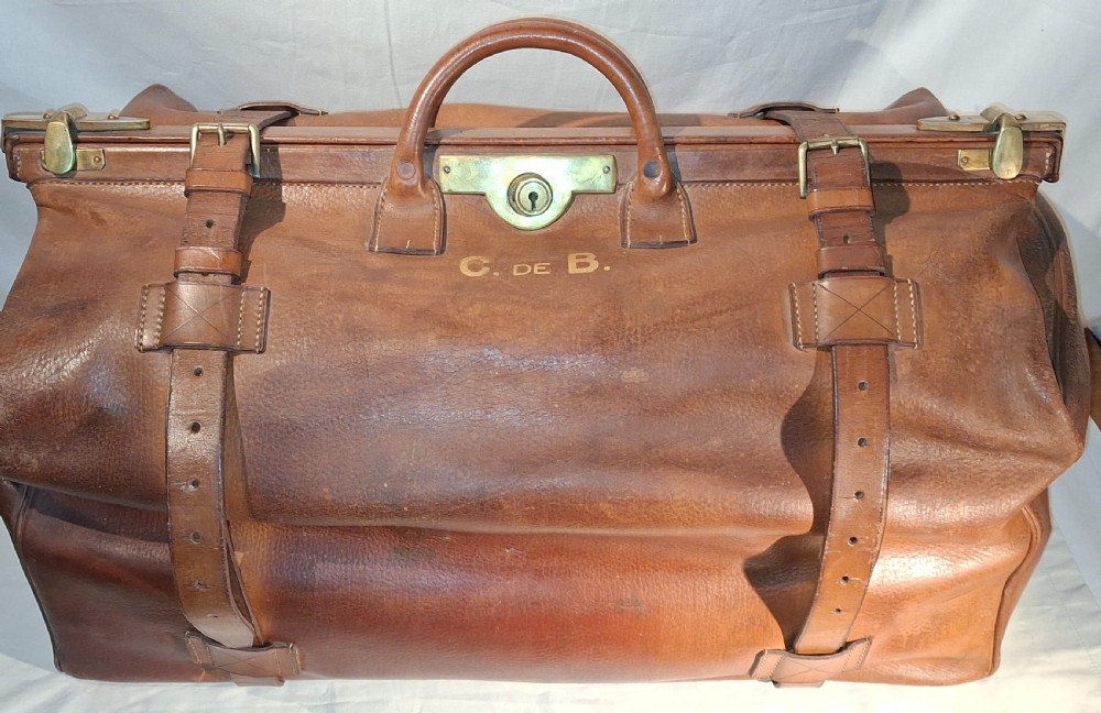A warm brown leather Gladstone bag featuring twin buckle straps, a brass central lock mechanism, a thick handle, and gold initials.