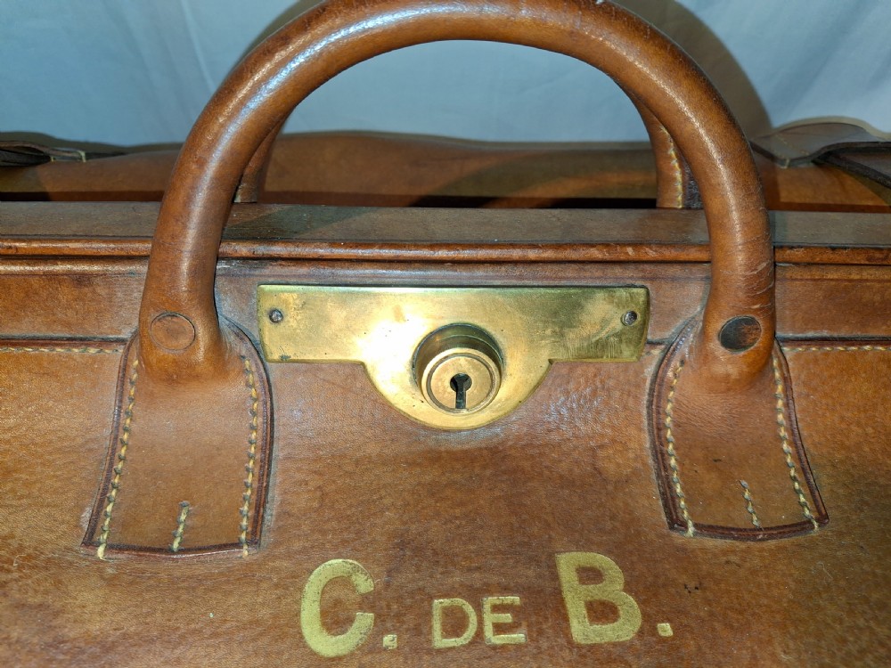 A warm brown leather Gladstone bag featuring twin buckle straps, a brass central lock mechanism, a thick handle, and gold initials. View VII