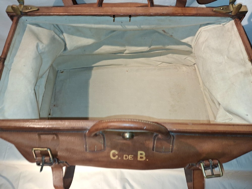 A warm brown leather Gladstone bag featuring twin buckle straps, a brass central lock mechanism, a thick handle, and gold initials. View VIII