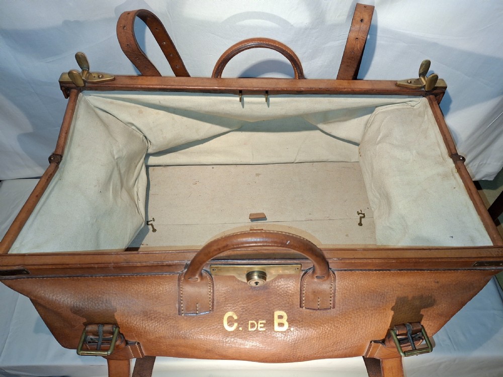A warm brown leather Gladstone bag featuring twin buckle straps, a brass central lock mechanism, a thick handle, and gold initials. View IX
