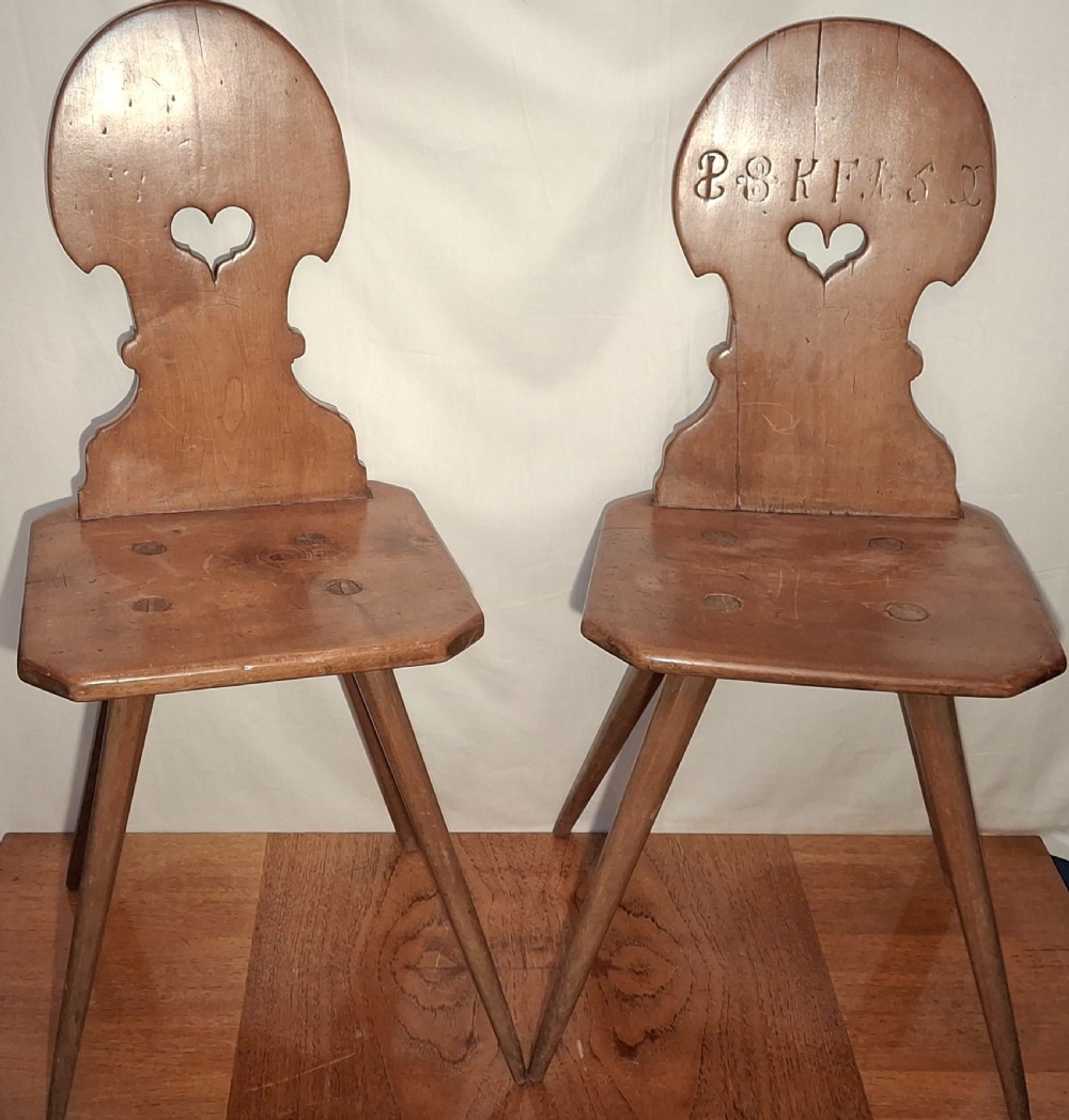 A matching pair of warm brown carved wooden Tyrolean side chairs featuring heart-shaped backrest cutouts, carved inscriptions, and splayed legs.