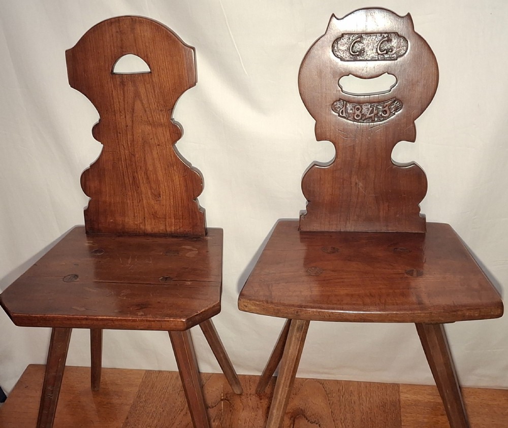 A pair of carved brown wooden Tyrolean side chairs featuring splayed legs, shaped backrests, and etched 1843 date details.