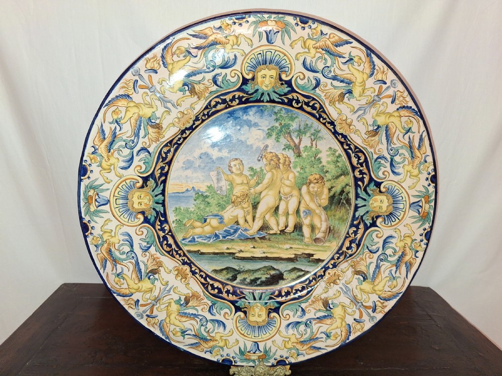 A circular 19th-century tin-glazed majolica charger featuring central putti in a landscape surrounded by grotesque border scrollwork in cobalt blue and yellow.