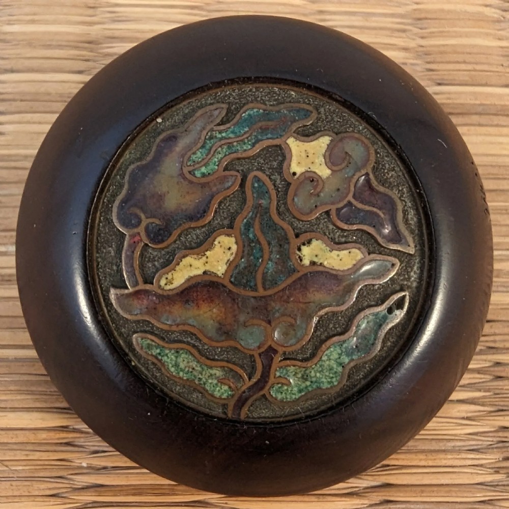 A circular dark wood Japanese kagamibuta netsuke with a central inset metal plate featuring green, yellow, and purple champleve enamel.