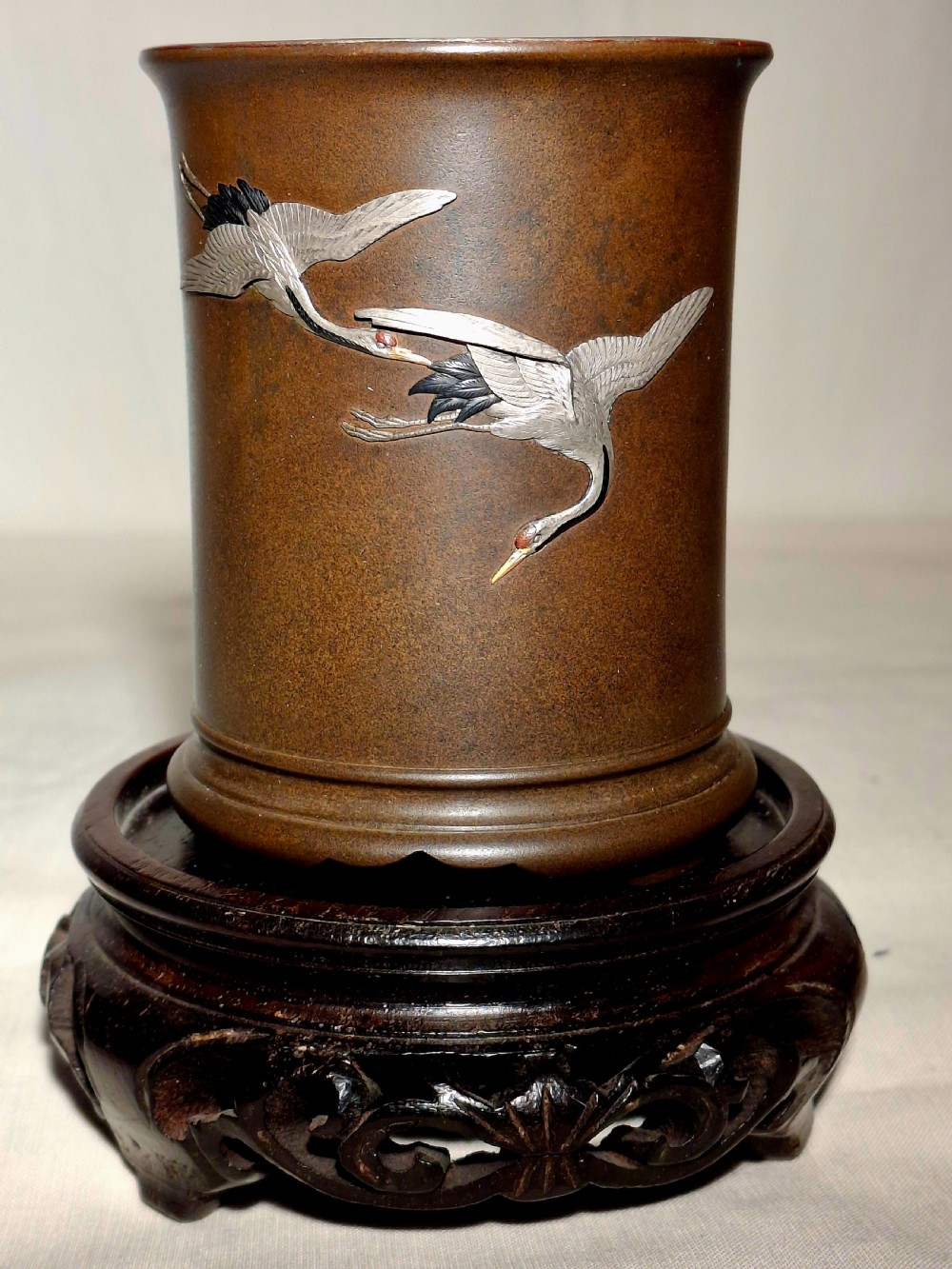 A dark brown cylindrical bronze brush pot decorated with inlaid silver cranes in flight, resting on a carved dark wooden base.
