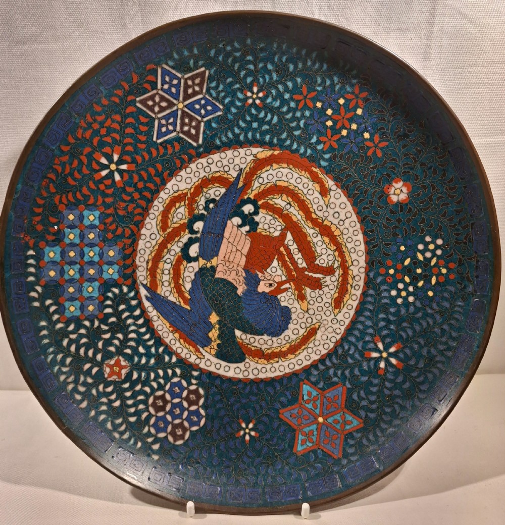 A circular Japanese cloisonne enamel charger depicting a central blue and red phoenix surrounded by intricate geometric and floral patterns on a dark teal field.