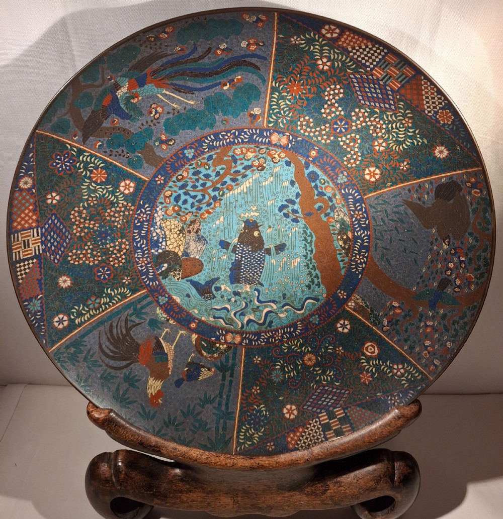 A large circular Japanese cloisonne enamel charger featuring a central leaping koi carp framed by segmented floral and bird panels on a dark carved wooden stand.