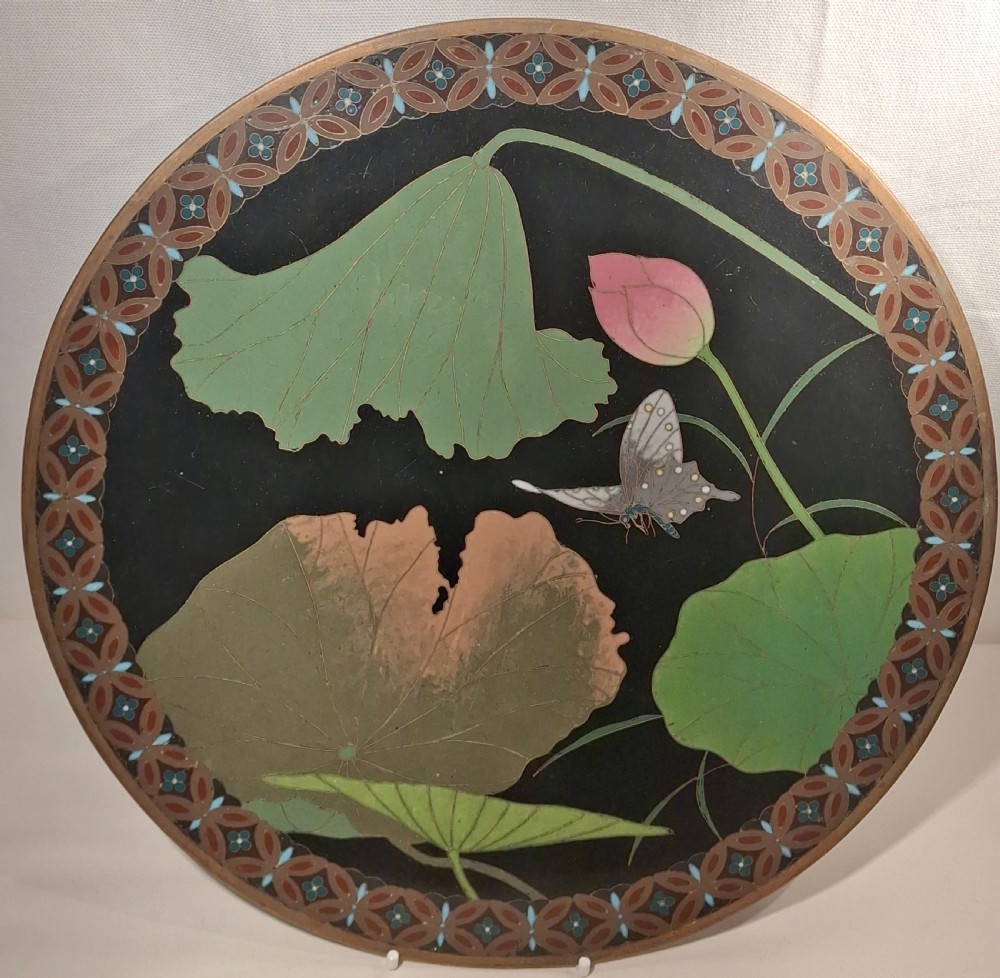 A circular Japanese cloisonne enamel plaque featuring green lotus leaves, a pink flower, and a butterfly on a black background framed by a geometric border.