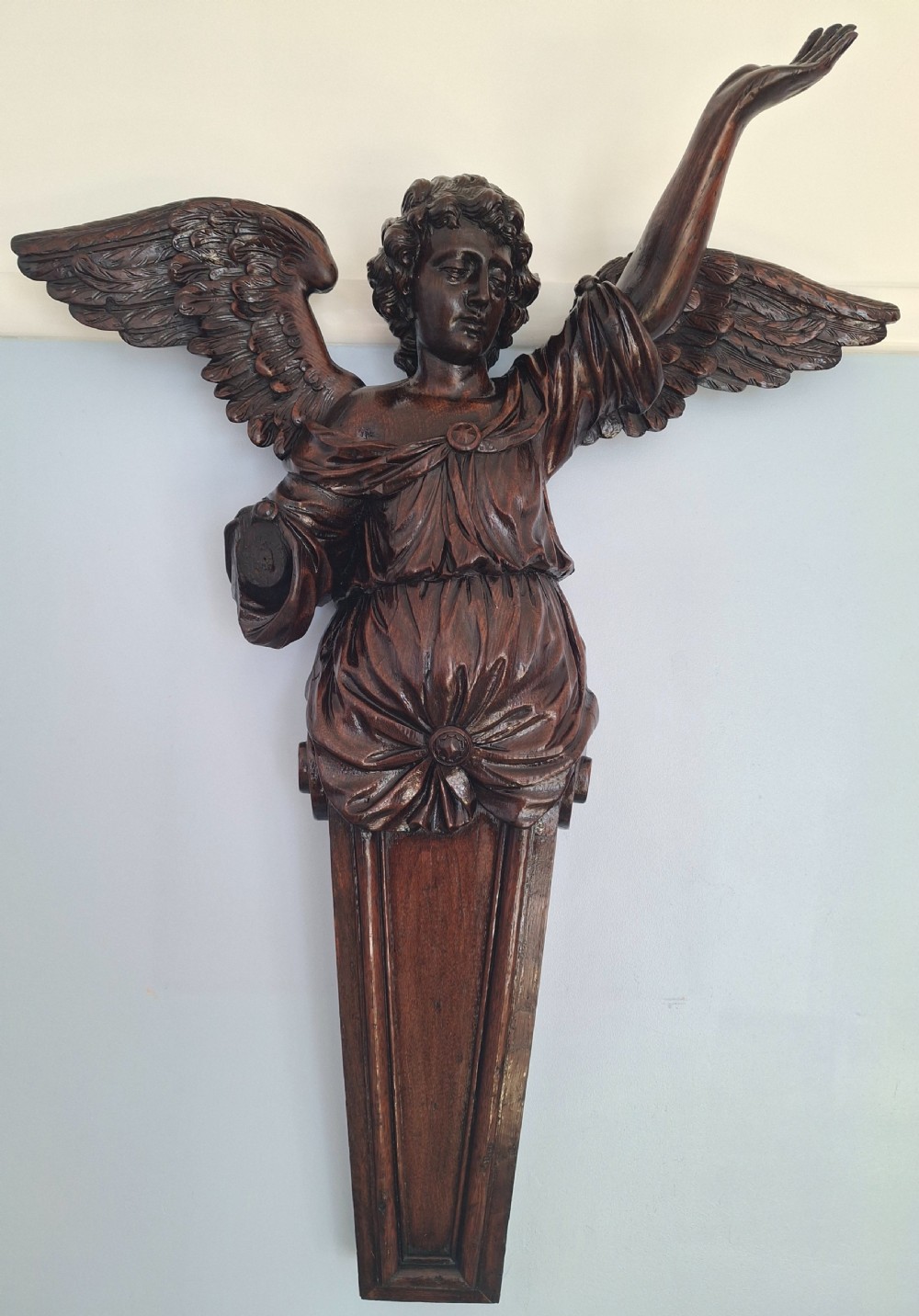 A dark carved oak wall sculpture depicting a winged angel figure with an outstretched arm resting on a tapered panel base.