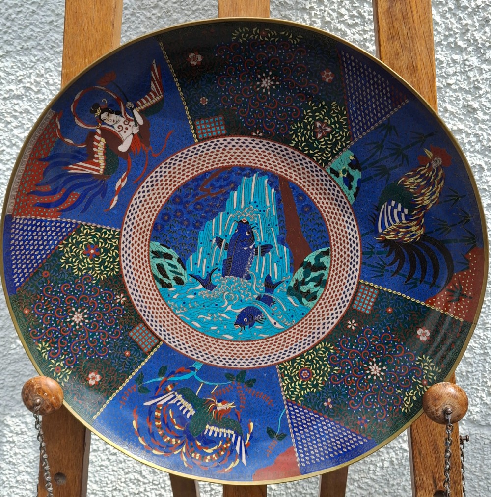 A large circular Japanese cloisonne enamel charger depicting a leaping blue koi carp in a central medallion surrounded by decorative patterned reserves.