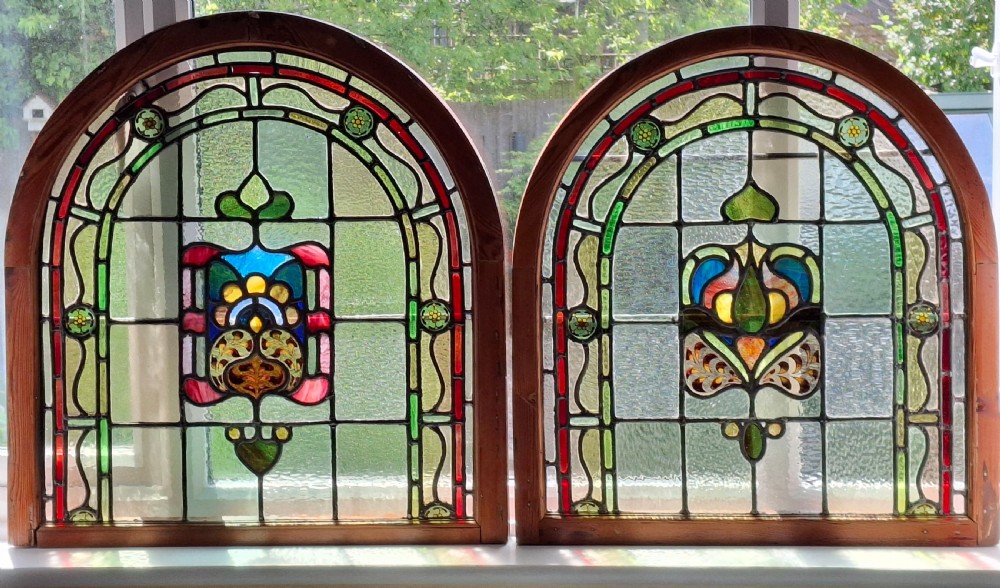 A pair of arched Edwardian stained glass windows set in timber frames featuring central Art Nouveau stylised floral motifs in red, blue, green, and yellow.