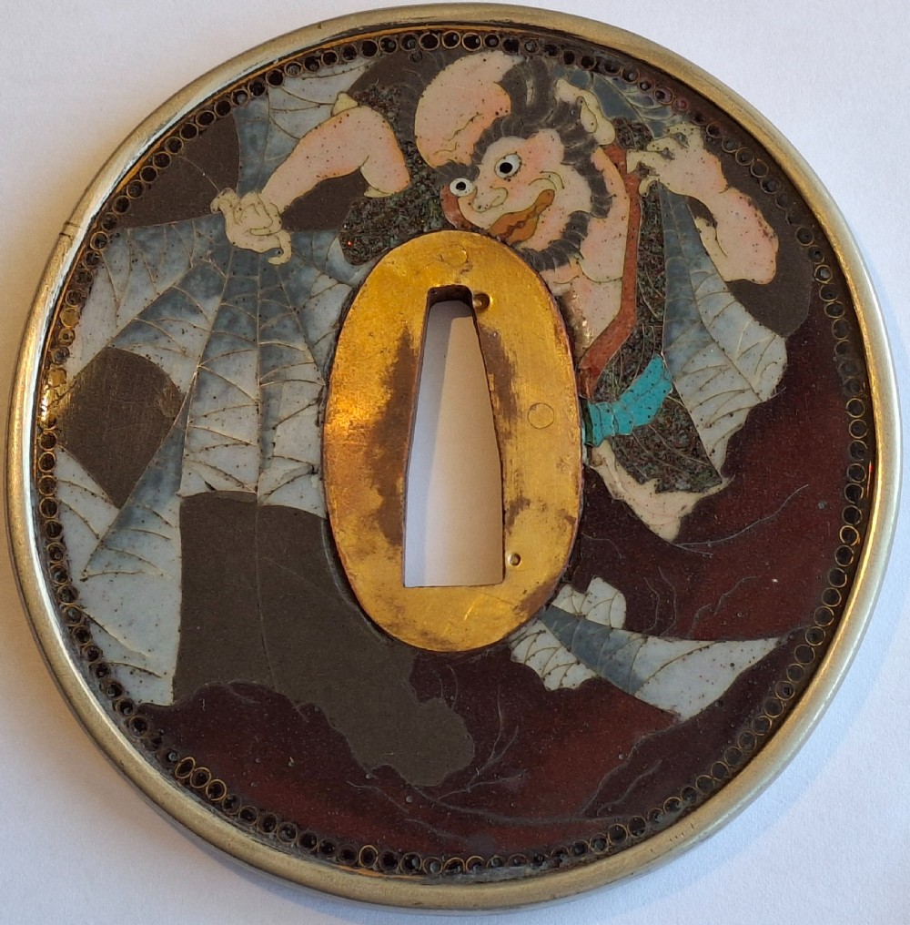 A circular Japanese cloisonne enamel tsuba featuring an intricate figure from folklore set against a dark ground with a central gilt opening.