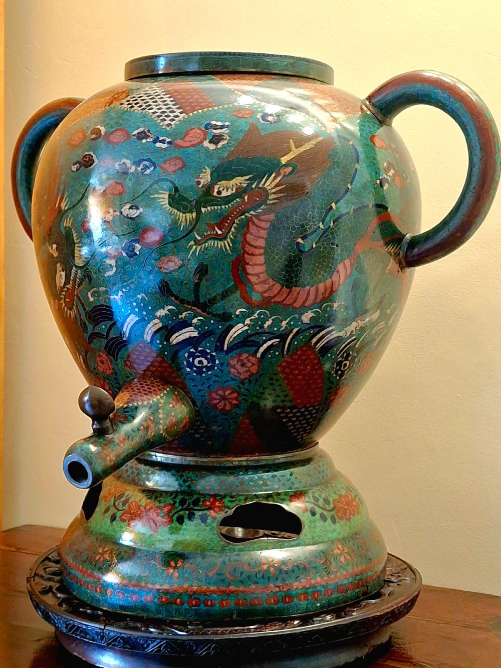 A large Meiji period Japanese cloisonne enamel water dispenser decorated with a dragon above sea waves, featuring twin handles, a lower spout, and a pedestal base.