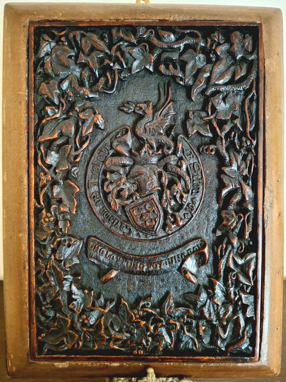 A dark brown carved wooden plaque featuring a detailed heraldic coat of arms with a dragon crest, framed by relief ivy leaves and a motto scroll.