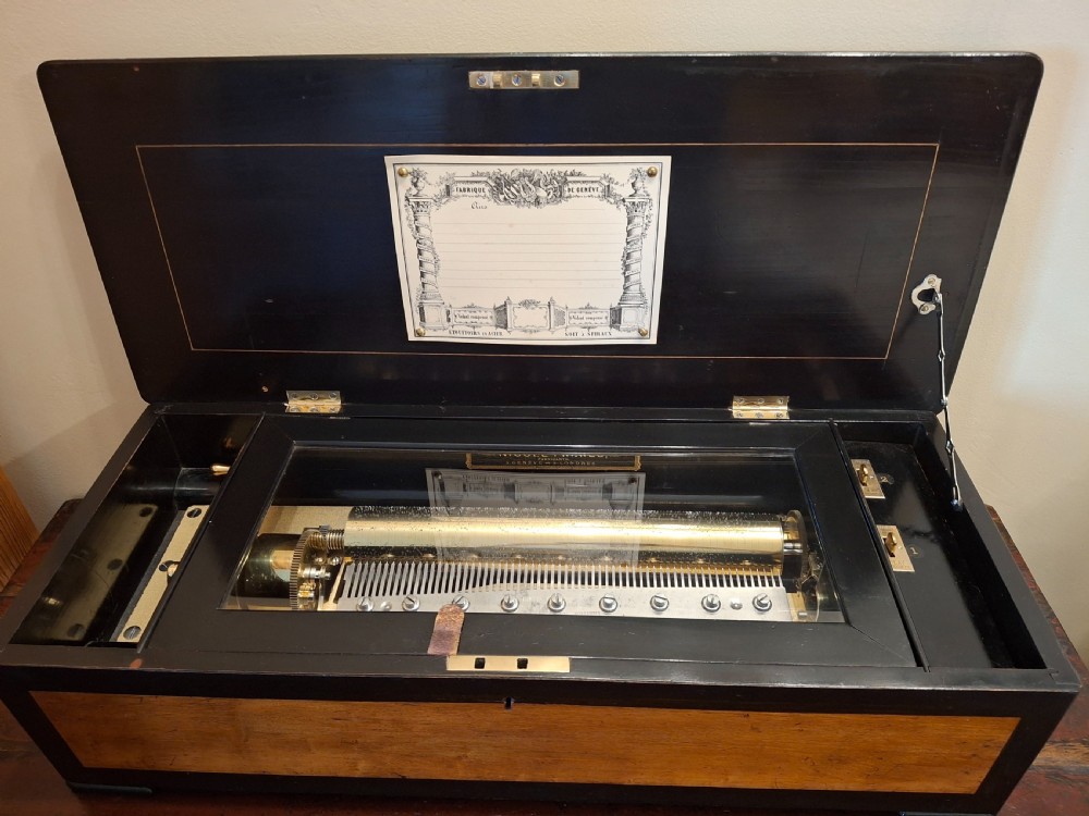 An opened ebonised wooden cylinder music box displaying a brass cylinder mechanism, internal glass cover, and original tune sheet attached under the lid.