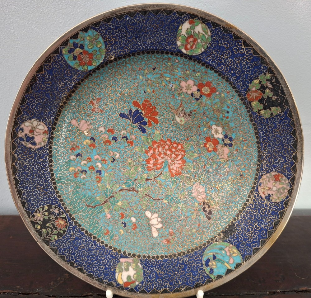 A circular Japanese Meiji period cloisonne enamel dish featuring a turquoise central section decorated with flowers and birds surrounded by a dark blue border.