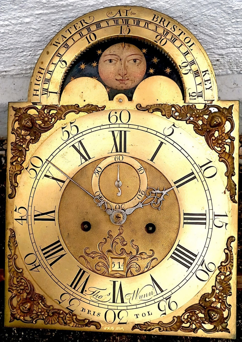 An 18th-century brass arched longcase clock dial featuring an integrated moonphase display, tidal indicator ring, and intricate engraved details.