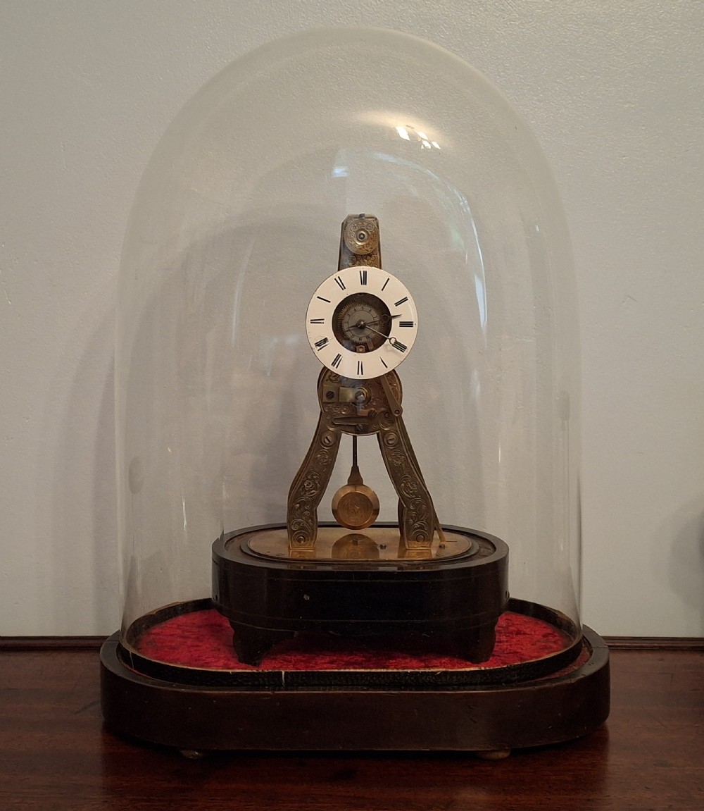 A miniature French brass skeleton clock with an exposed mechanical movement, white porcelain Roman numeral dial, and pendulum, housed beneath a clear glass dome on a dark wooden base.