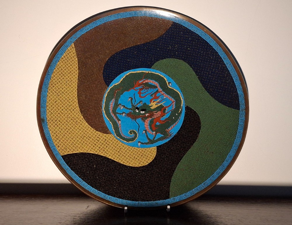 A circular Japanese cloisonne enamel plate featuring a central green dragon motif framed by swirling patterned segments in blue, brown, yellow, and green.