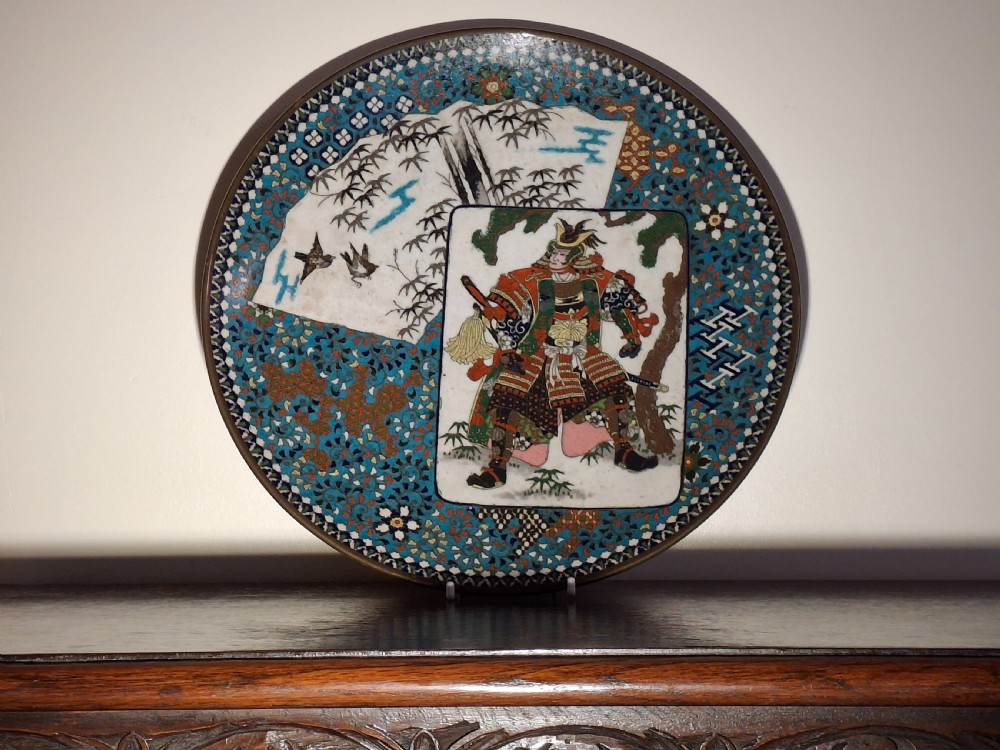 A circular Japanese cloisonne enamel plate featuring a central rectangular panel depicting a samurai warrior in full armour alongside a fan-shaped panel with bamboo foliage, set against a turquoise geometric pattern background.