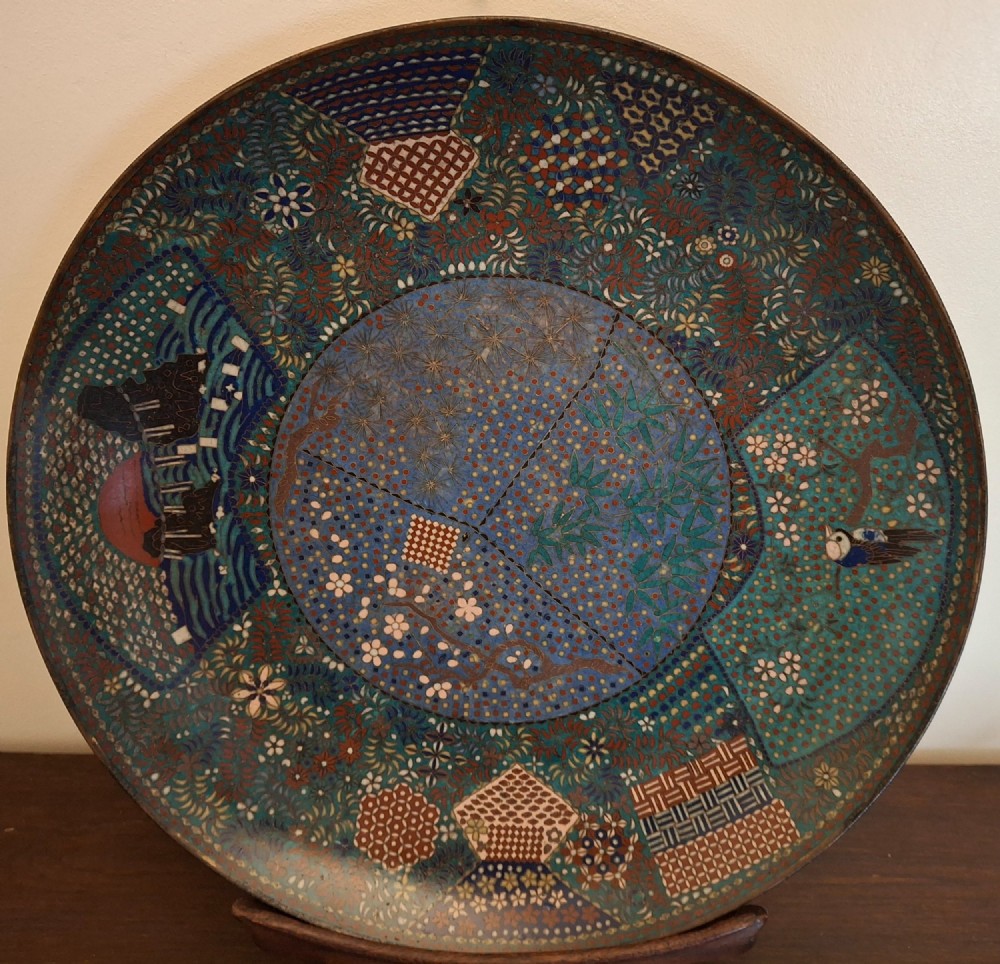 A circular Japanese cloisonne enamel charger decorated with geometric reserves, floral patterns, foliage, and bird motifs in blue, green, and terracotta tones.