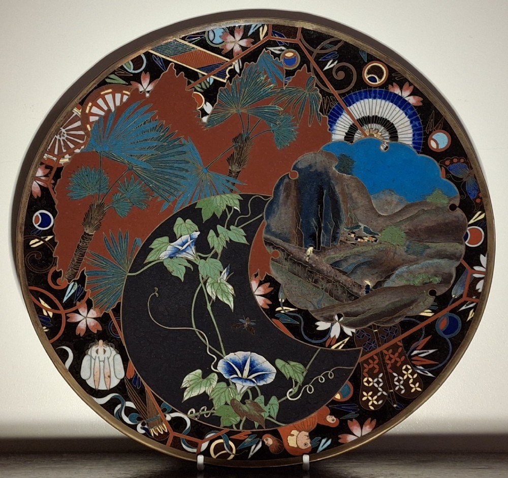 A circular Japanese cloisonne enamel plate featuring intricate floral, landscape, and geometric designs in blue, green, and red against a dark ground.