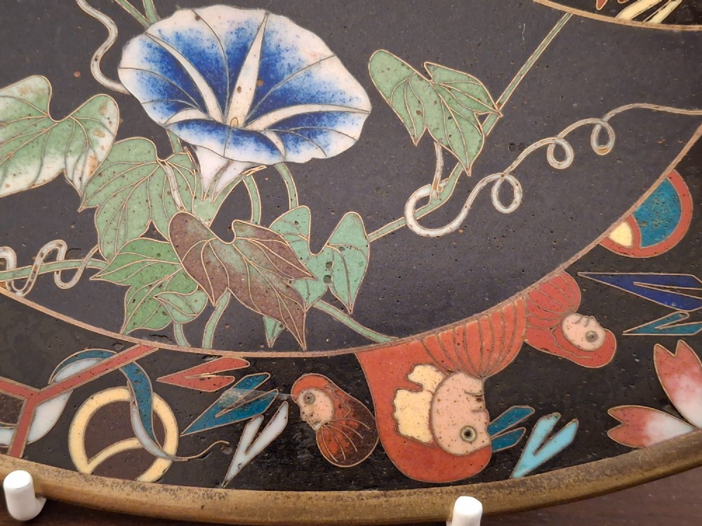 A circular Japanese cloisonne enamel plate featuring intricate floral, landscape, and geometric designs in blue, green, and red against a dark ground. View VI