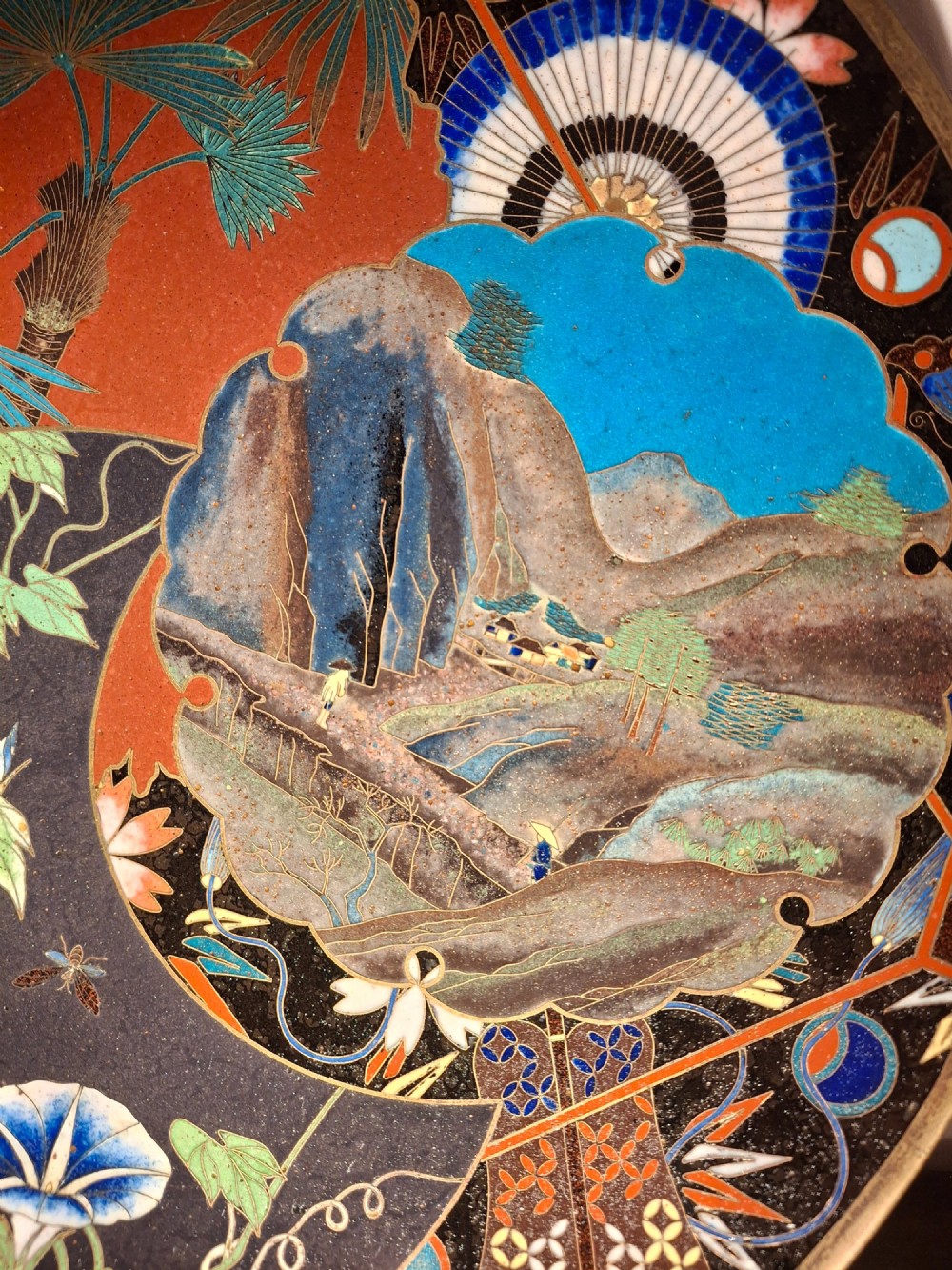 A circular Japanese cloisonne enamel plate featuring intricate floral, landscape, and geometric designs in blue, green, and red against a dark ground. View XI
