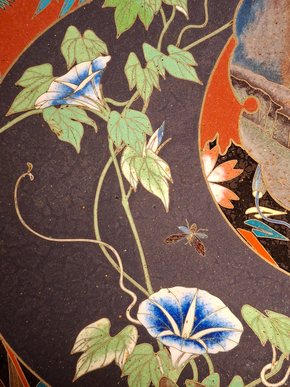 A circular Japanese cloisonne enamel plate featuring intricate floral, landscape, and geometric designs in blue, green, and red against a dark ground. View XII