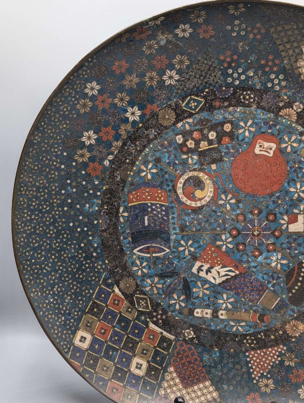 A circular Japanese cloisonne charger featuring a central blue medallion with a red Daruma figure surrounded by detailed geometric and floral patterned borders. View V