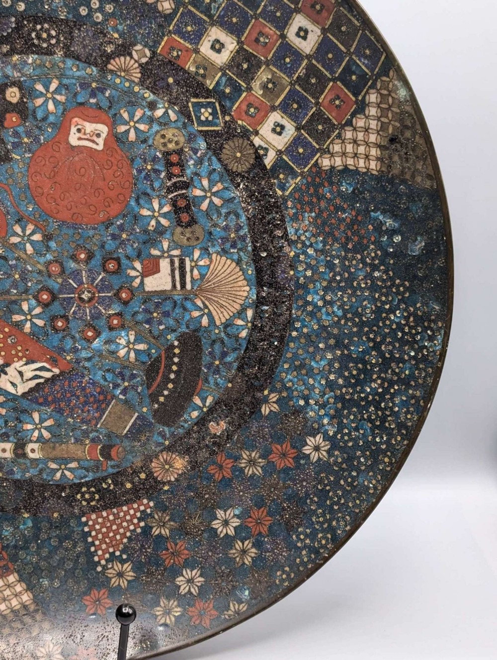 A circular Japanese cloisonne charger featuring a central blue medallion with a red Daruma figure surrounded by detailed geometric and floral patterned borders. View VI