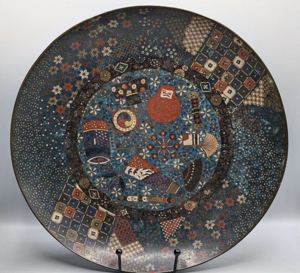 A circular Japanese cloisonne charger featuring a central blue medallion with a red Daruma figure surrounded by detailed geometric and floral patterned borders.