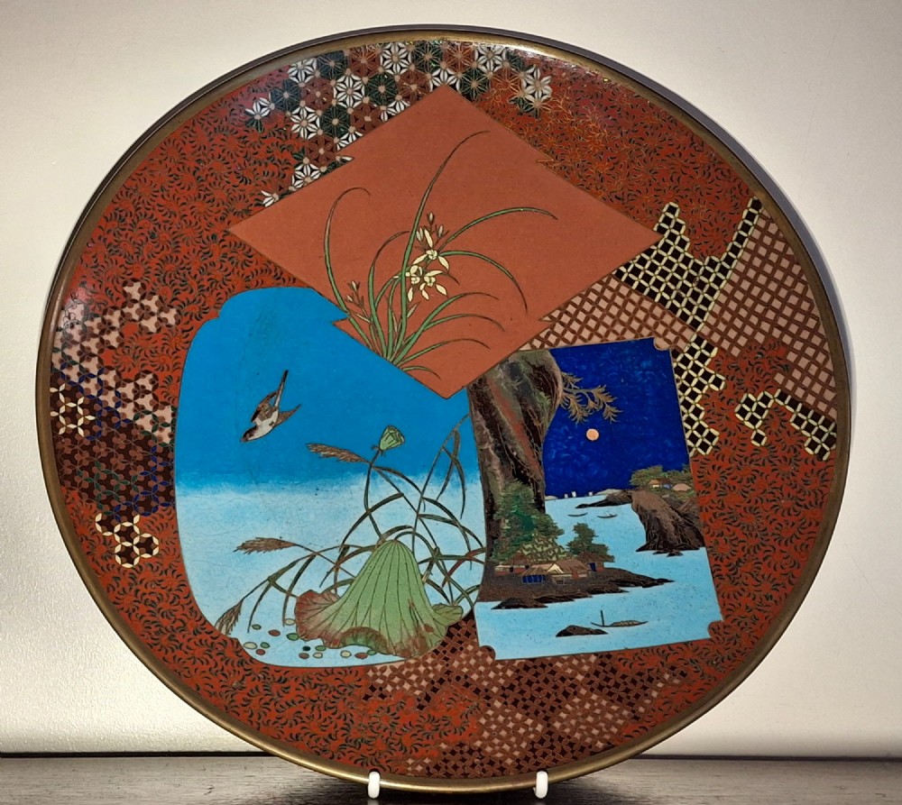 A circular Japanese cloisonne enamel plate featuring geometric patterned borders around asymmetrical panels depicting flora, a bird, and a nocturnal coastal scene.