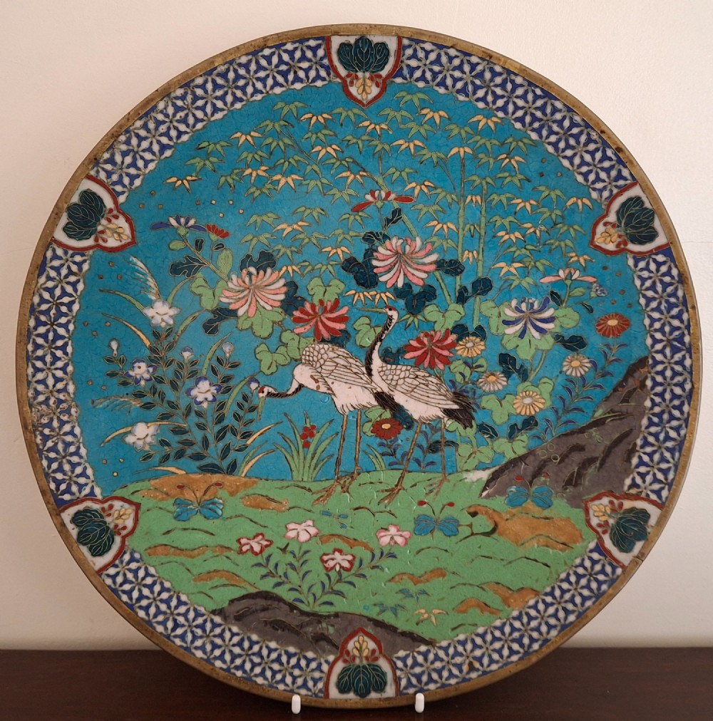 A circular Japanese cloisonne enamel decorative plate decorated with two wading birds surrounded by flowers against a turquoise ground with an ornamental geometric border.