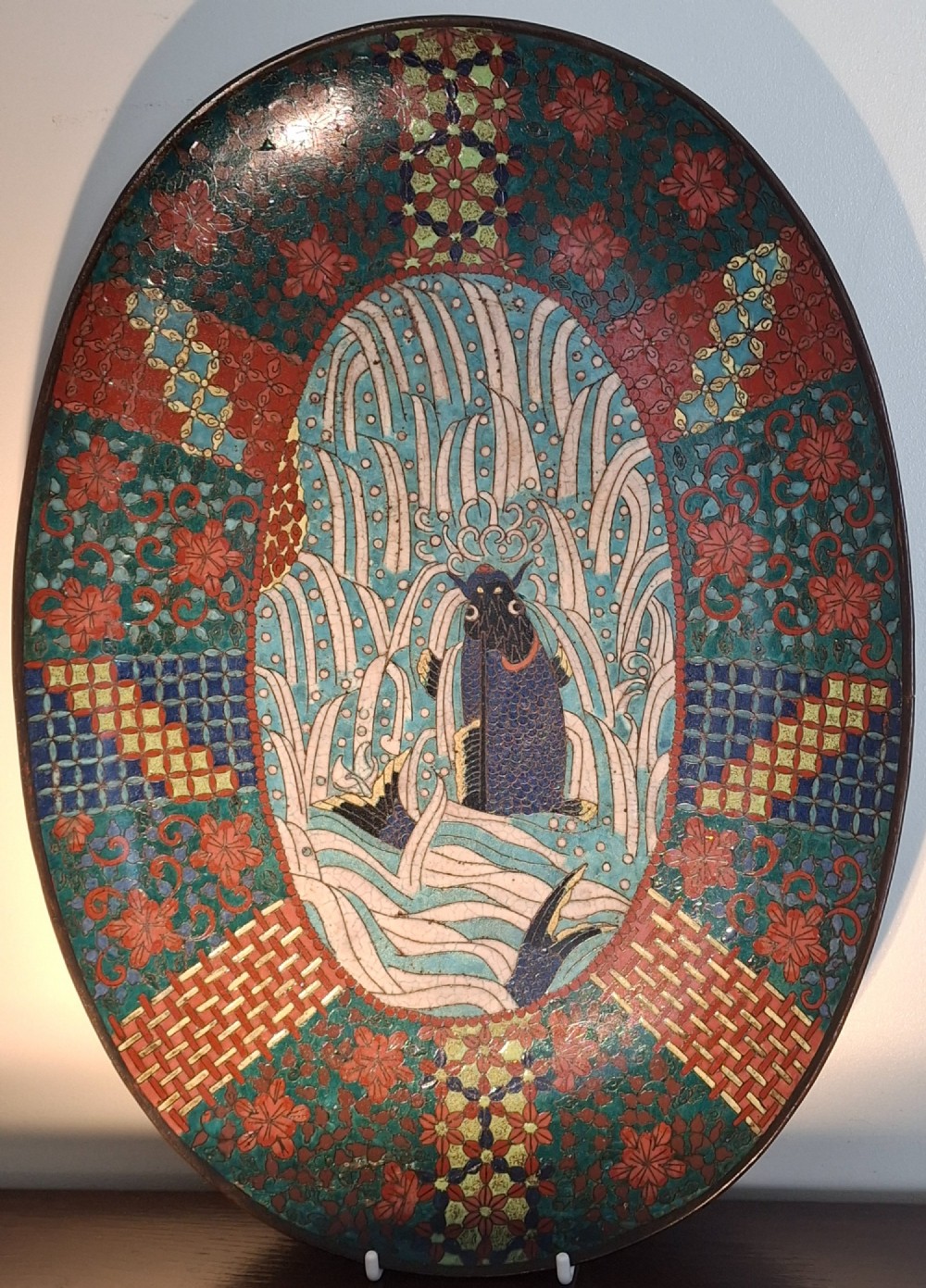 An oval Japanese cloisonne enamel tray displaying a central blue leaping koi carp within stylized waves, surrounded by geometric and red floral patterned borders.