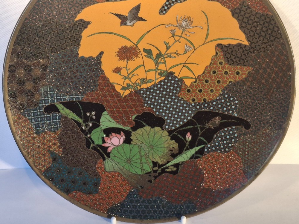 A circular Japanese cloisonne enamel dish featuring a central yellow reserve with a bird and chrysanthemums alongside lotus flowers against geometric mosaic patterns. View II