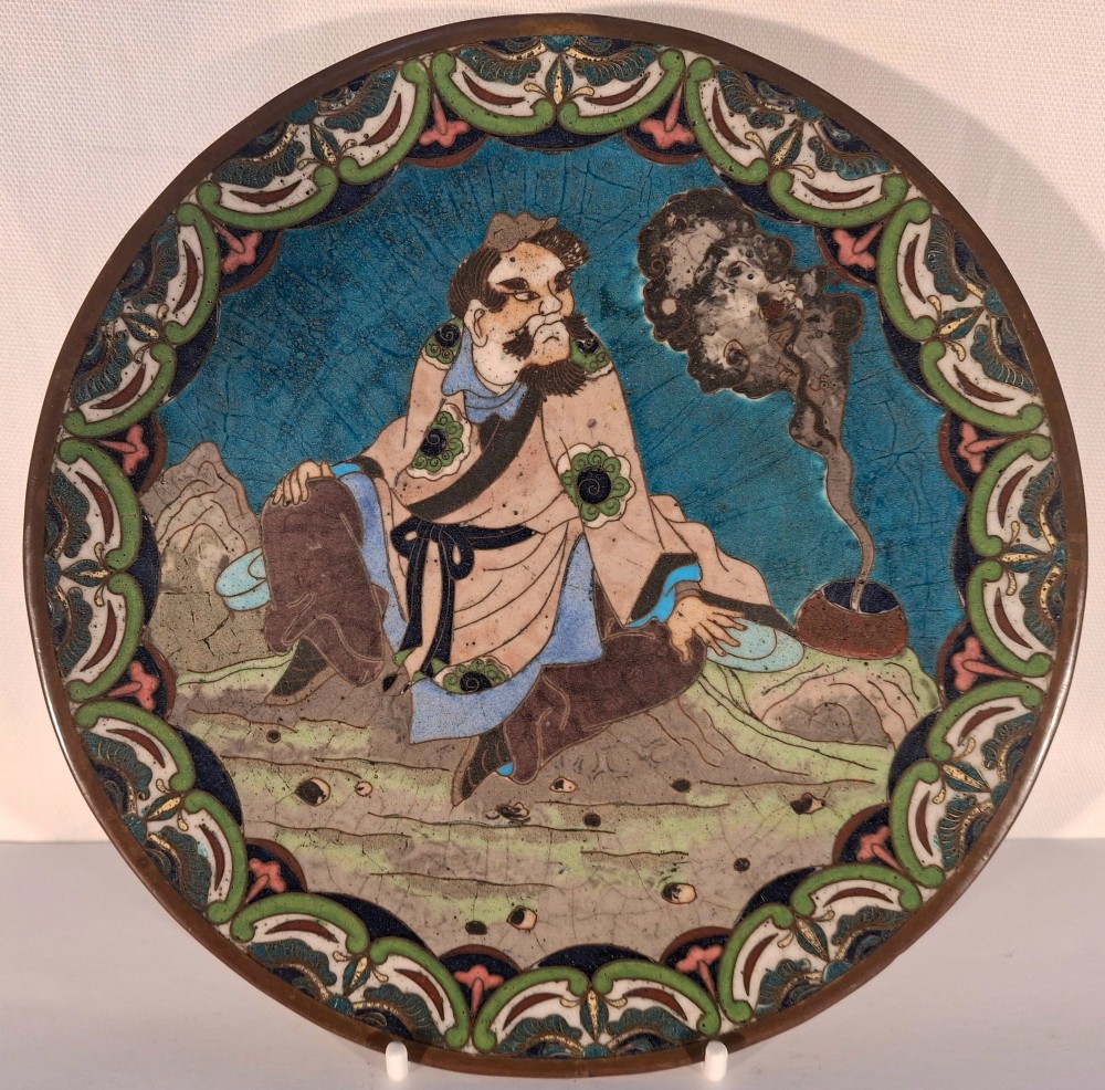 A circular Japanese cloisonne enamel plate depicting a bearded seated figure beside a smoking pot on a turquoise background with a floral border.