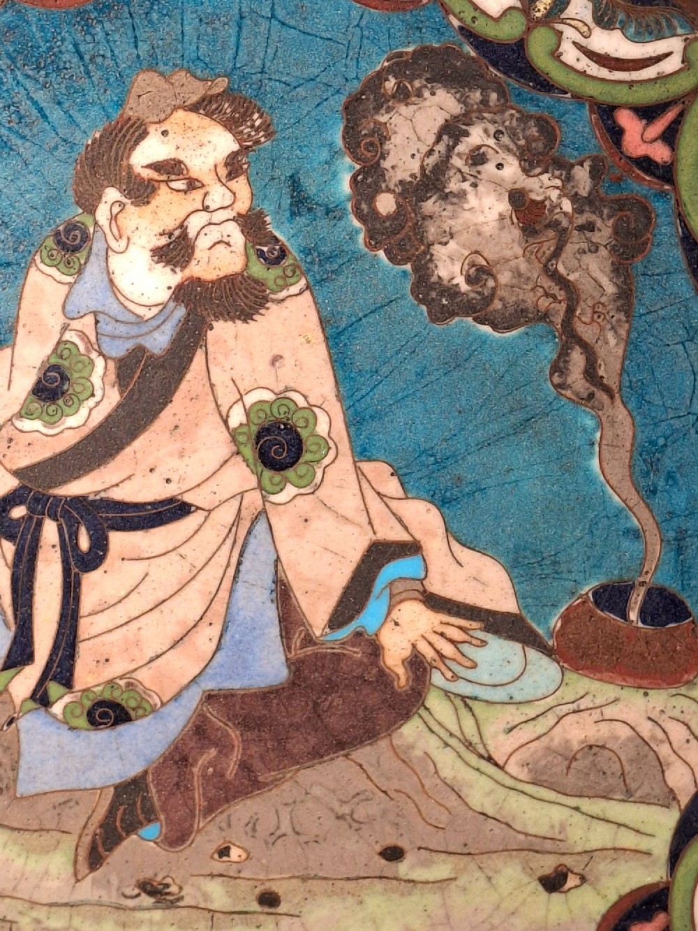A circular Japanese cloisonne enamel plate depicting a bearded seated figure beside a smoking pot on a turquoise background with a floral border. View III