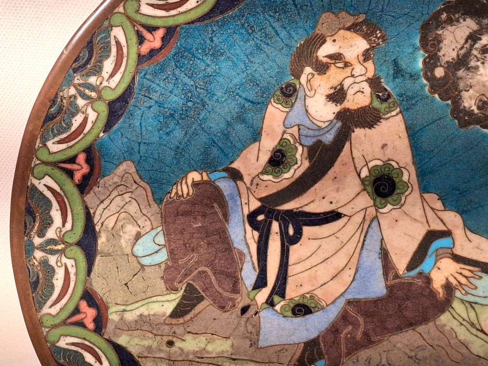 A circular Japanese cloisonne enamel plate depicting a bearded seated figure beside a smoking pot on a turquoise background with a floral border. View IV