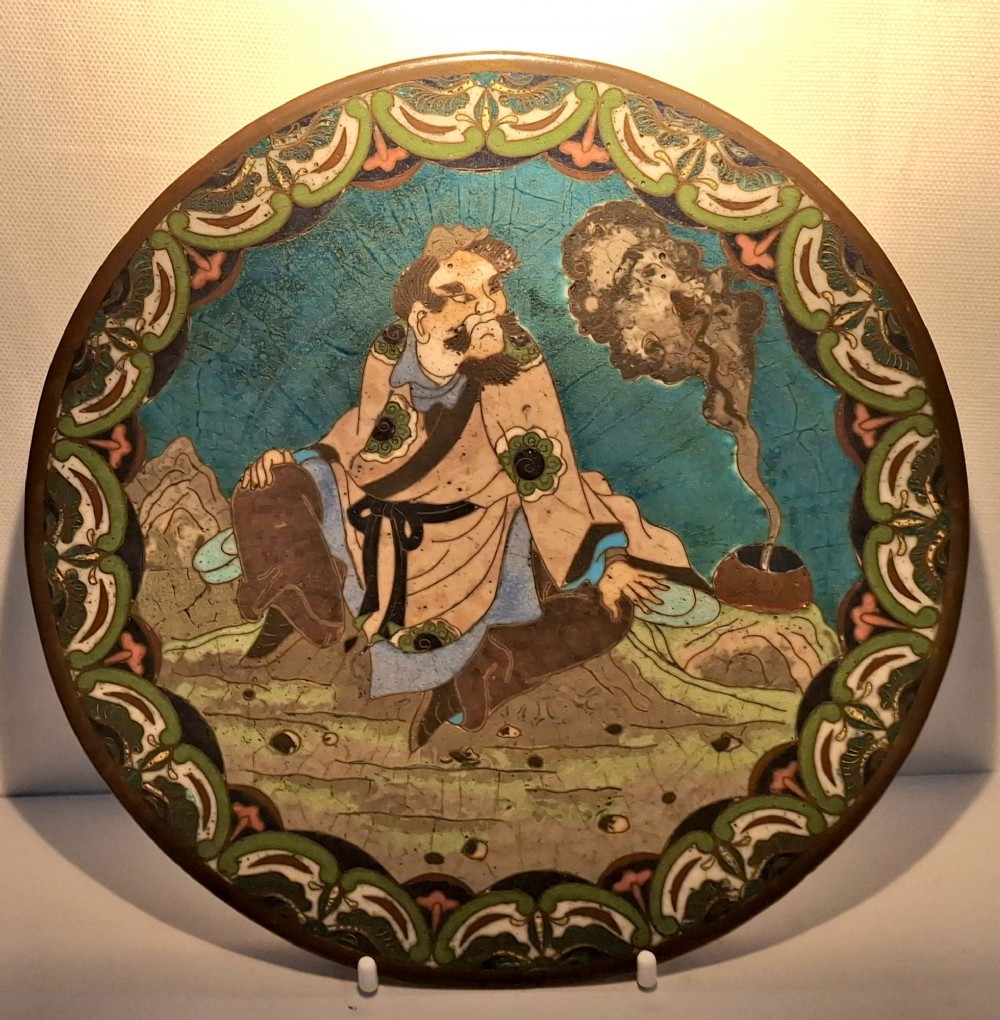 A circular Japanese cloisonne enamel plate depicting a bearded seated figure beside a smoking pot on a turquoise background with a floral border. View IX