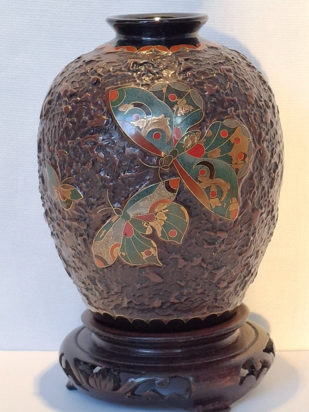 A bulbous dark brown ceramic vase with textured tree-bark surface and colourful enamel butterfly designs resting on a dark carved wooden stand. View II