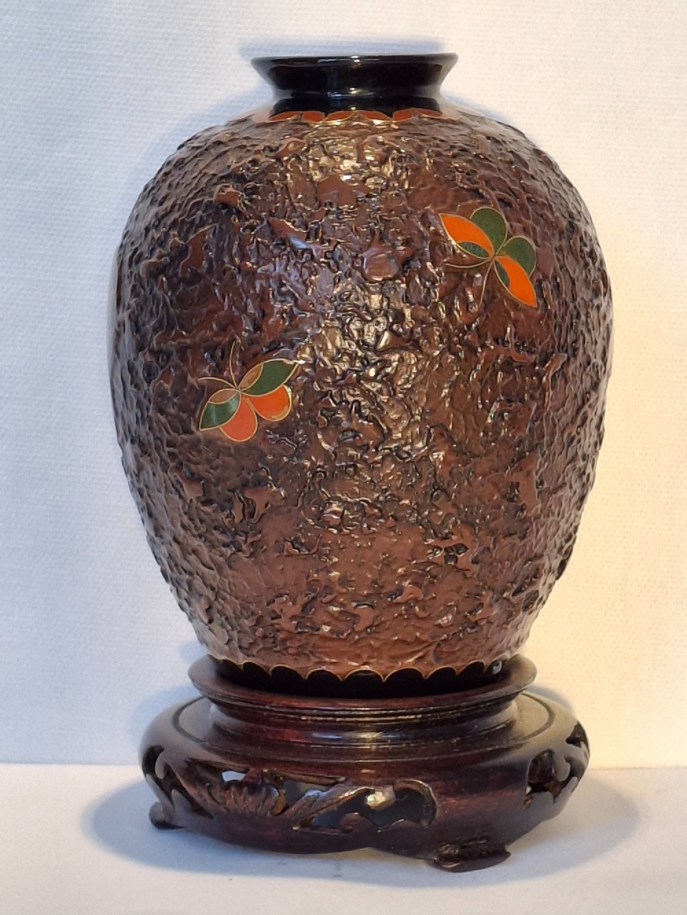 A bulbous dark brown ceramic vase with textured tree-bark surface and colourful enamel butterfly designs resting on a dark carved wooden stand. View IV