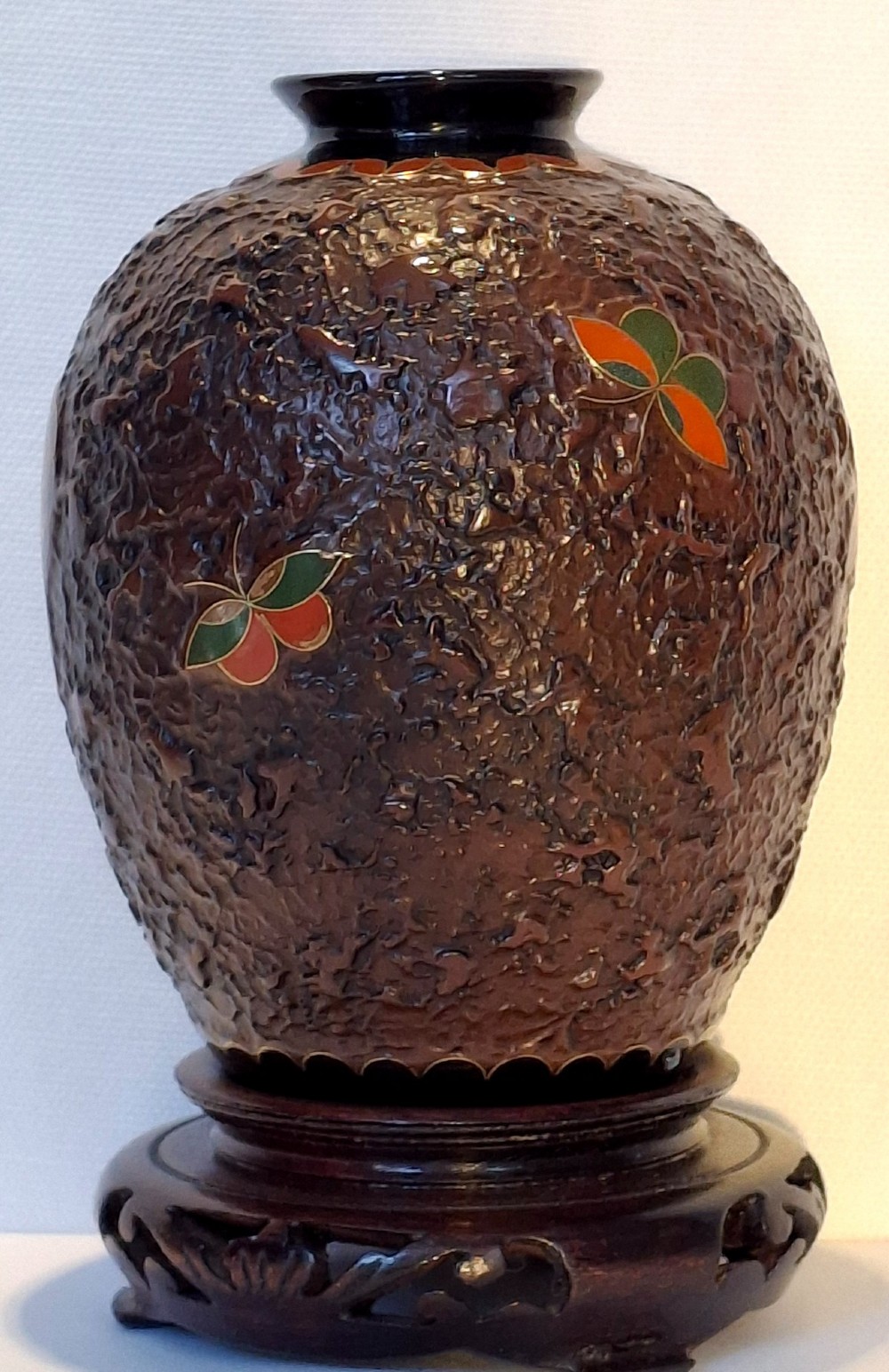 A bulbous dark brown ceramic vase with textured tree-bark surface and colourful enamel butterfly designs resting on a dark carved wooden stand. View V
