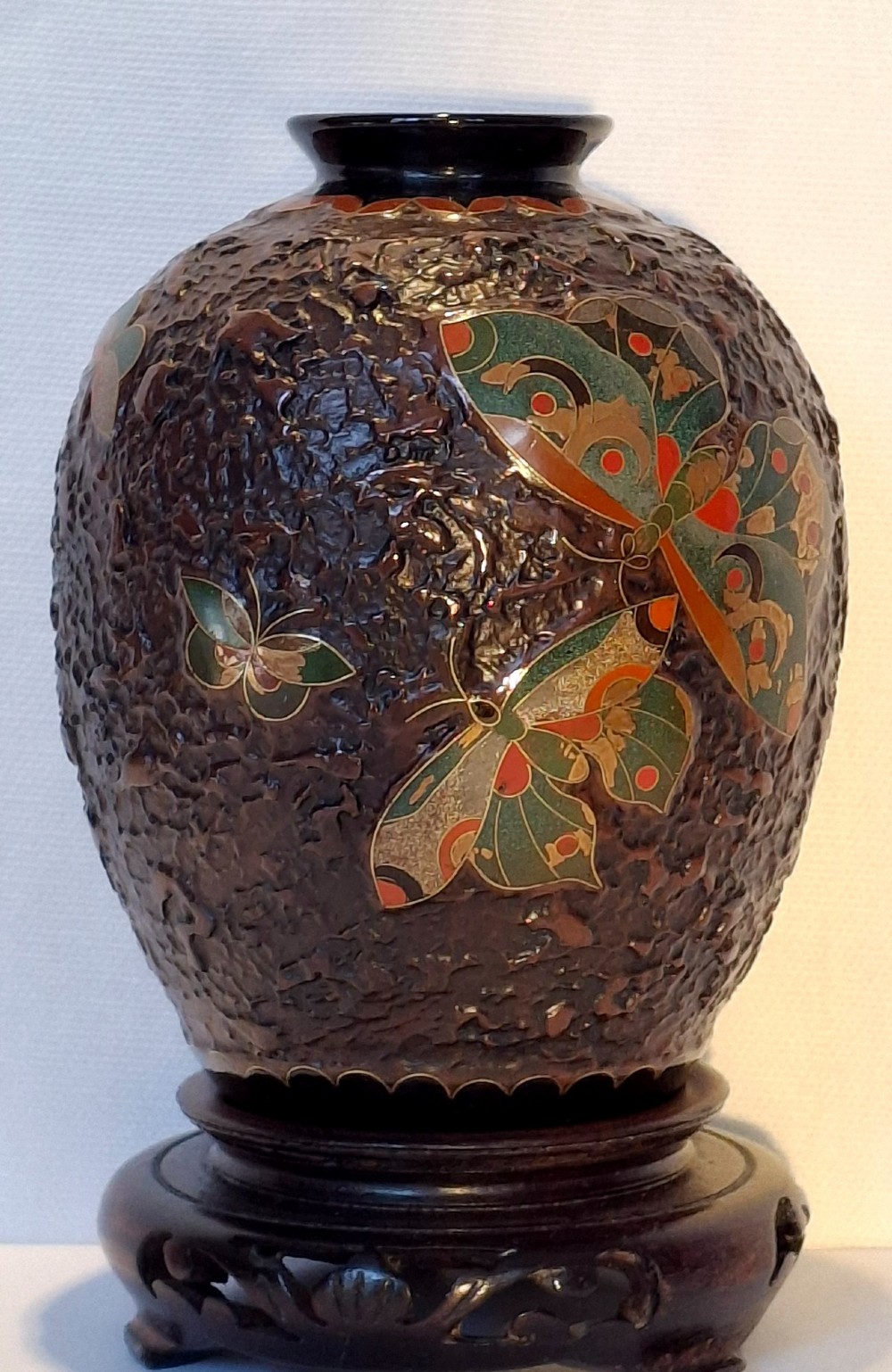 A bulbous dark brown ceramic vase with textured tree-bark surface and colourful enamel butterfly designs resting on a dark carved wooden stand. View VII