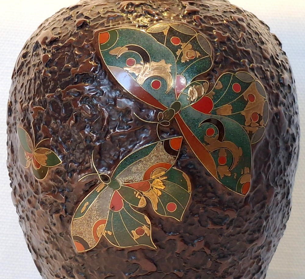 A bulbous dark brown ceramic vase with textured tree-bark surface and colourful enamel butterfly designs resting on a dark carved wooden stand. View VIII