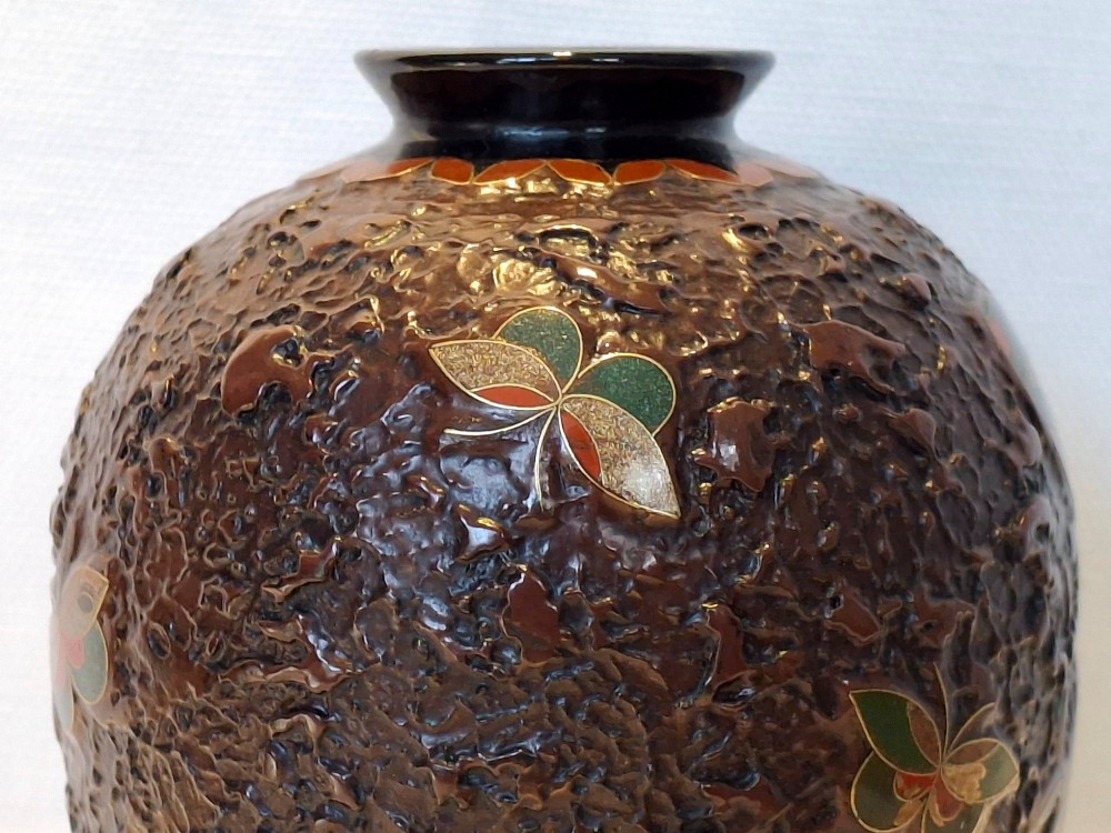 A bulbous dark brown ceramic vase with textured tree-bark surface and colourful enamel butterfly designs resting on a dark carved wooden stand. View X