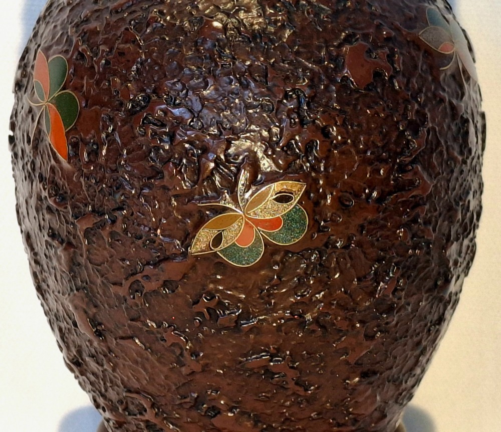 A bulbous dark brown ceramic vase with textured tree-bark surface and colourful enamel butterfly designs resting on a dark carved wooden stand. View XI