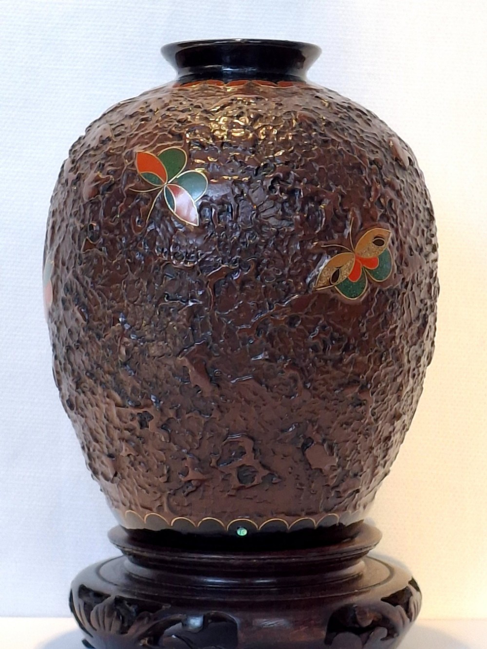 A bulbous dark brown ceramic vase with textured tree-bark surface and colourful enamel butterfly designs resting on a dark carved wooden stand. View XV